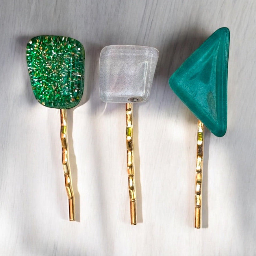 Three hair pins with gold metal bases and colorful glass tips on a light wooden surface.