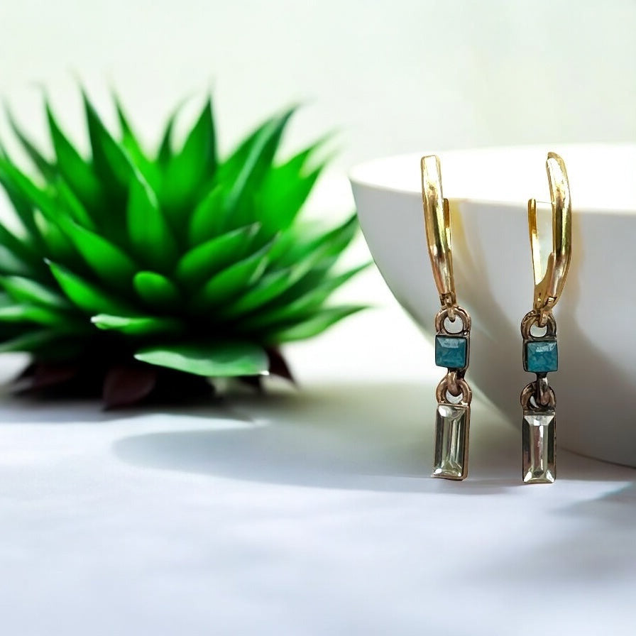 Gold earrings with green gemstones on a white surface with a green plant in the background
