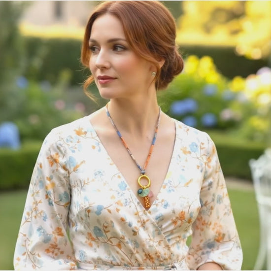 Woman in a floral dress wearing a fabric cord and glass charms necklace with orange and gold speckled pendant, pale green ginkgo leaf, and pink and blue glass accents, in a garden setting. Handmade by Eclectic Blue Studio, Edinburgh.