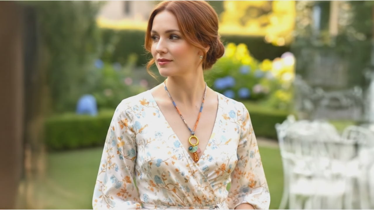 Woman in a floral dress wearing a fabric cord and glass charms necklace with orange and gold speckled pendant, pale green ginkgo leaf, and pink and blue glass accents, in a garden setting. Handmade by Eclectic Blue Studio, Edinburgh.